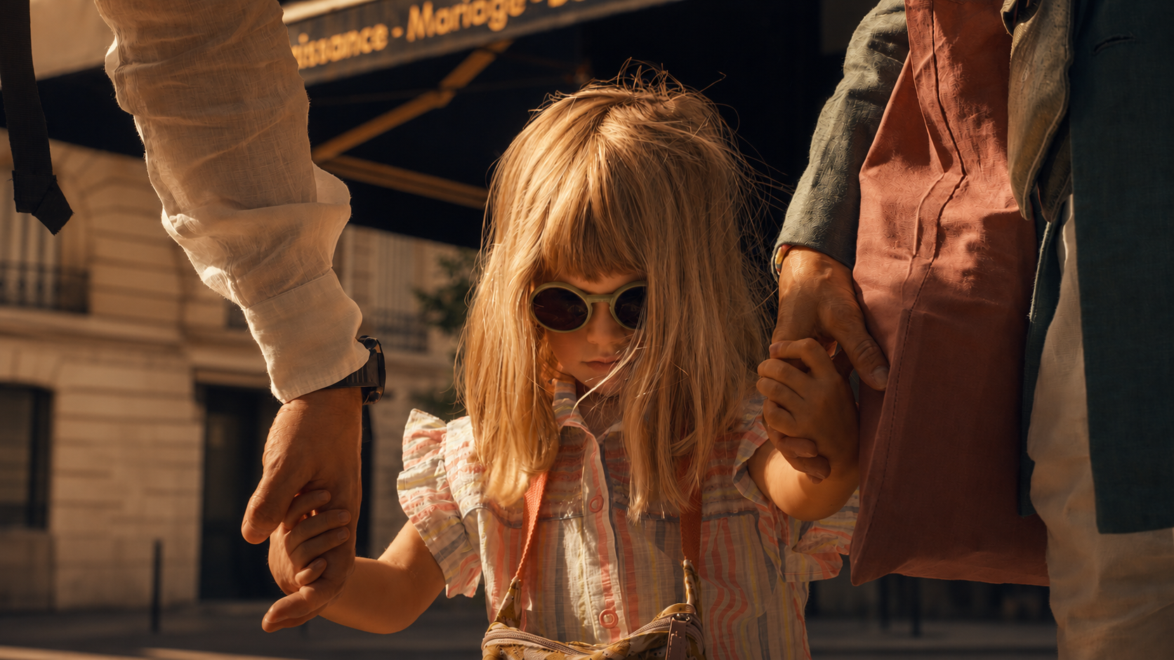 A young girl in sunglasses holding her parents' hands on a Paris street, documentary film still
